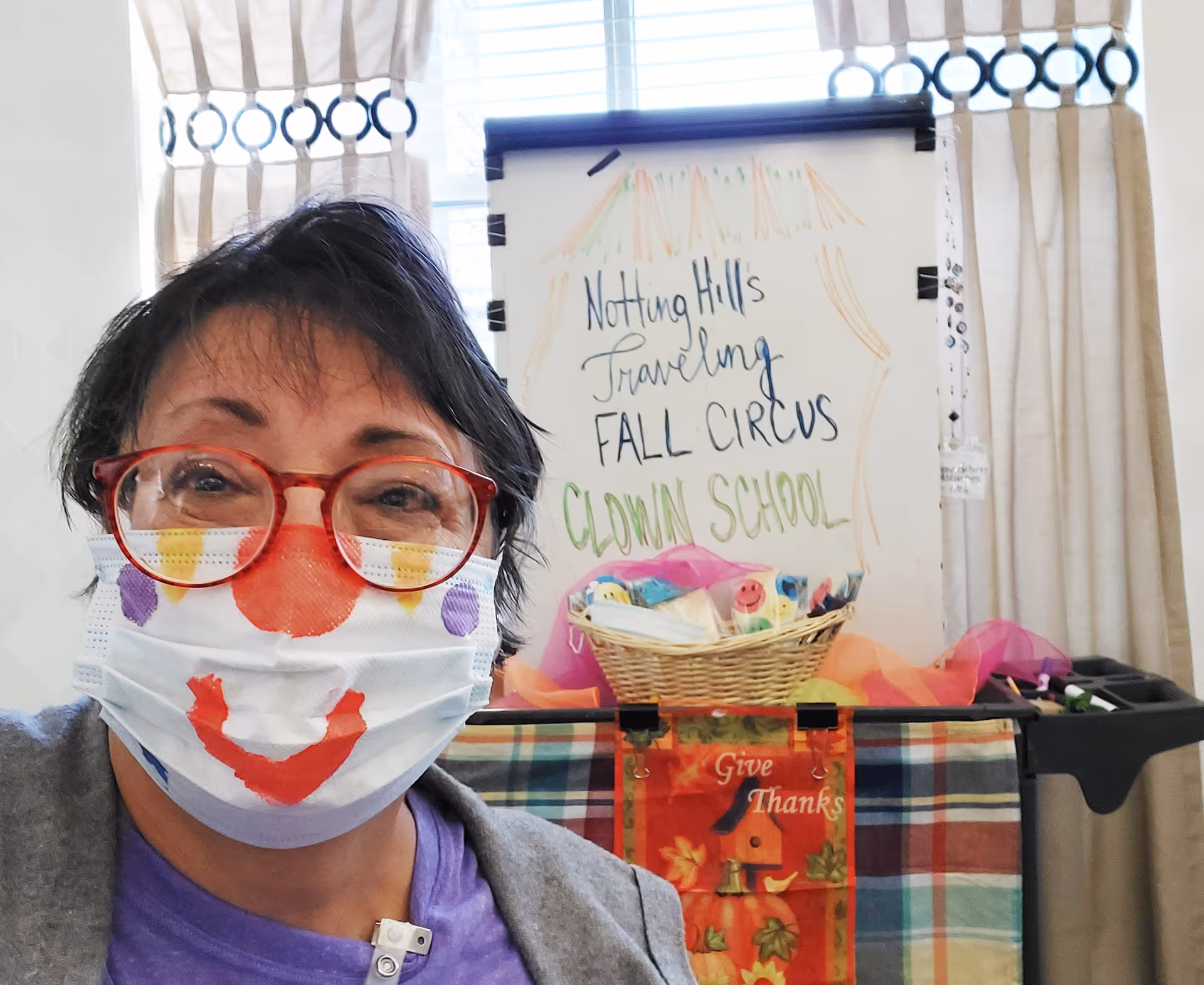 A person wearing red glasses and a face mask decorated with a clown face, standing indoors in front of a sign that reads 'Notting Hill's Traveling Fall Circus Clown School'. There is a basket with colorful items and a 'Give Thanks' decoration below the sign.