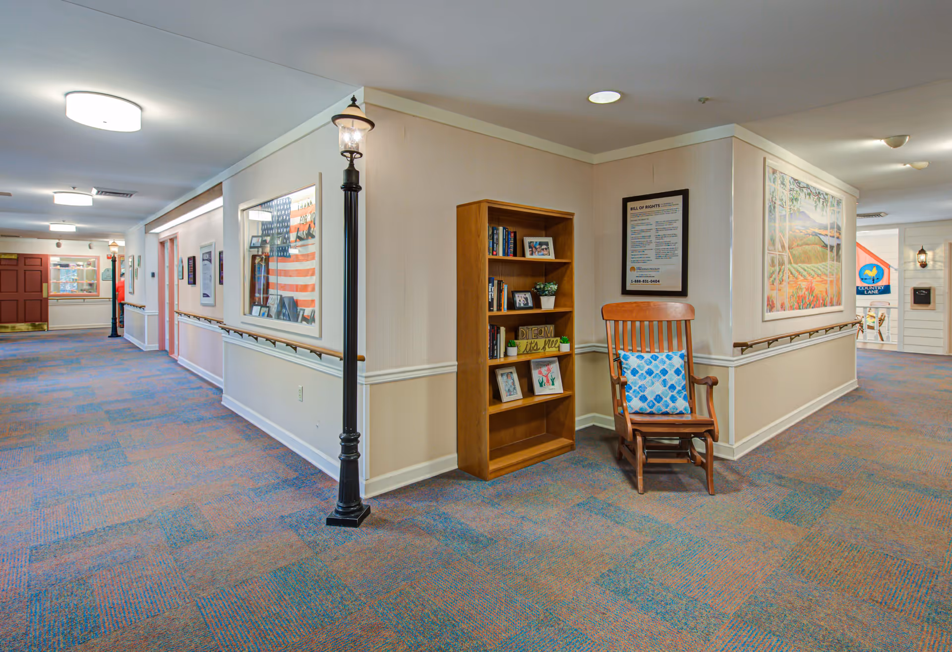 Carpeted interior hallway of a memory care facility with handrails, a bookshelf, a wooden rocking chair with a cushion, and decorative lampposts.
