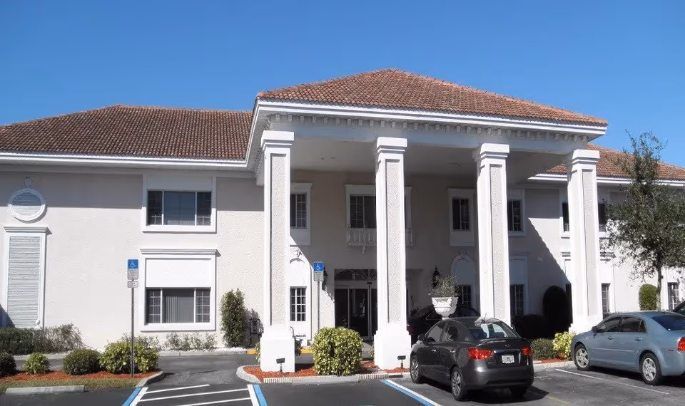 Front exterior view of a two-story senior living facility building with a tiled roof, large white columns at the entrance, several windows, and a parking lot with cars and handicap parking spaces in front.