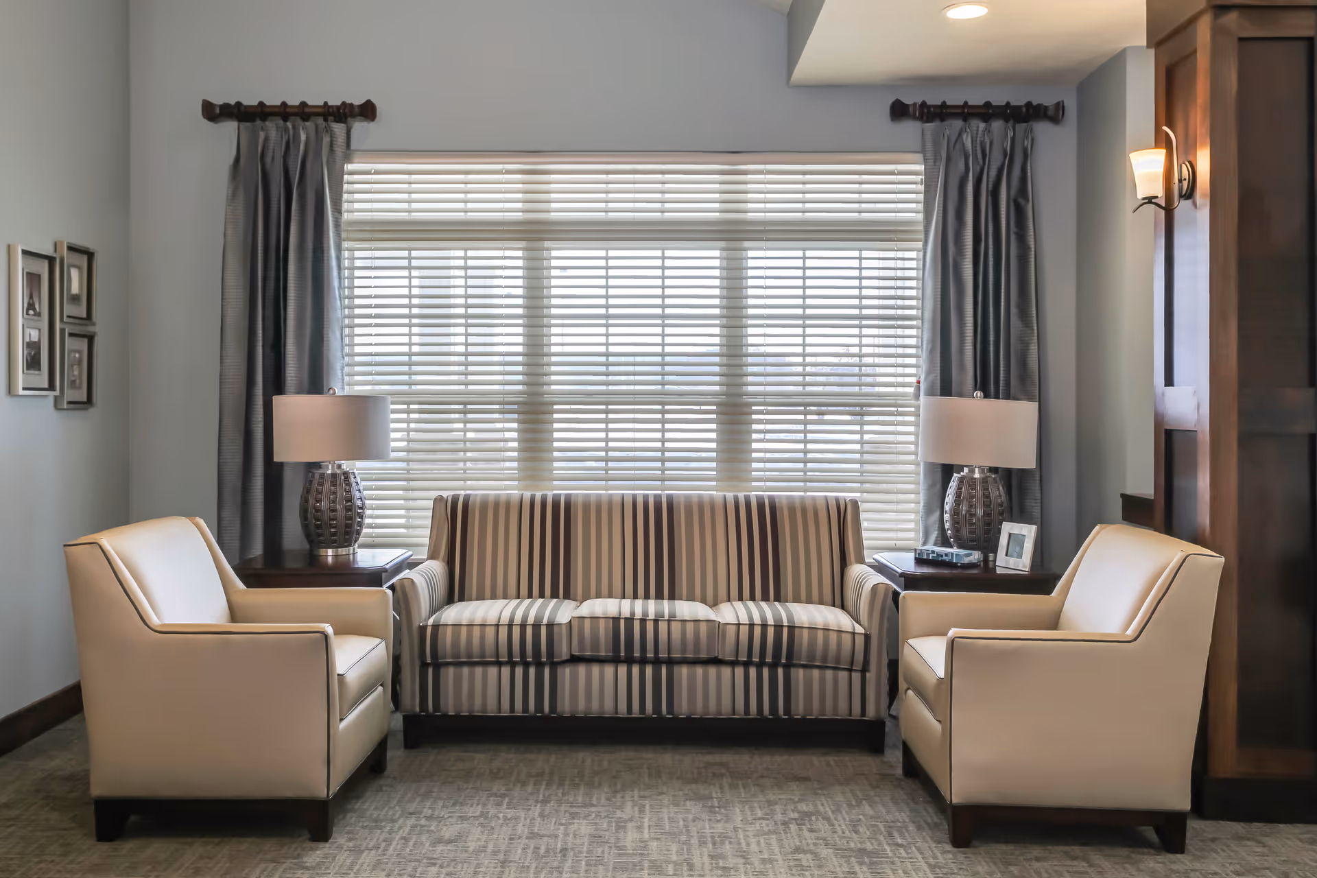 A cozy seating area with a striped sofa flanked by two armchairs, side tables and lamps in front of a large window with blinds.