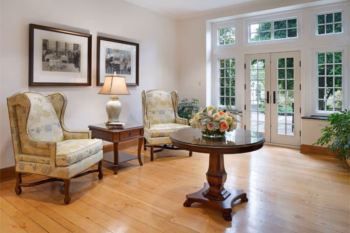 A cozy sitting area with two upholstered armchairs, a wooden side table with a lamp and books, and a round wooden table with a floral arrangement. The room has light-colored walls, wooden flooring, framed artwork on the wall, and large windows and glass doors letting in natural light with a view of greenery outside.