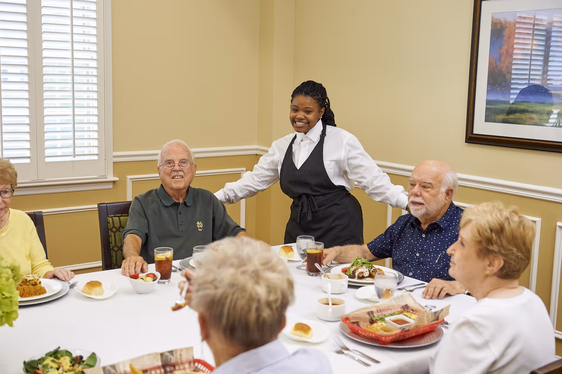A group of elderly people seated around a dining table enjoying a meal together in a well-lit room with beige walls and a window with white shutters. A smiling waitress in a white shirt and black apron stands behind them with her arms outstretched.