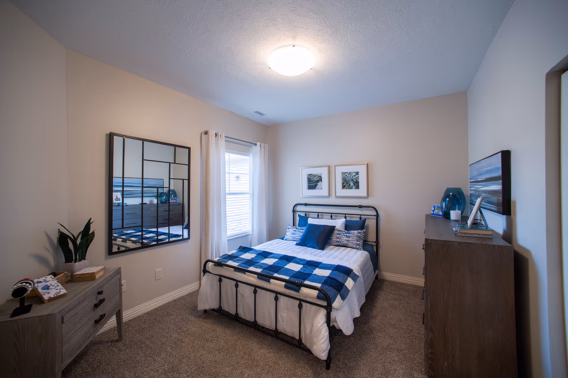 Neatly furnished bedroom with a metal-framed bed dressed in blue-and-white bedding, a dresser, decorative mirror, and a window with curtains.