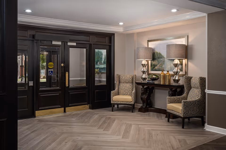 An elegant interior seating area near a set of dark wooden double doors with glass panels. The seating area features two patterned armchairs with beige cushions, a dark wooden console table with decorative lion-shaped legs, two modern table lamps with geometric bases, and decorative items including gold vases and a framed landscape painting on the wall. The floor has a light wood herringbone pattern.