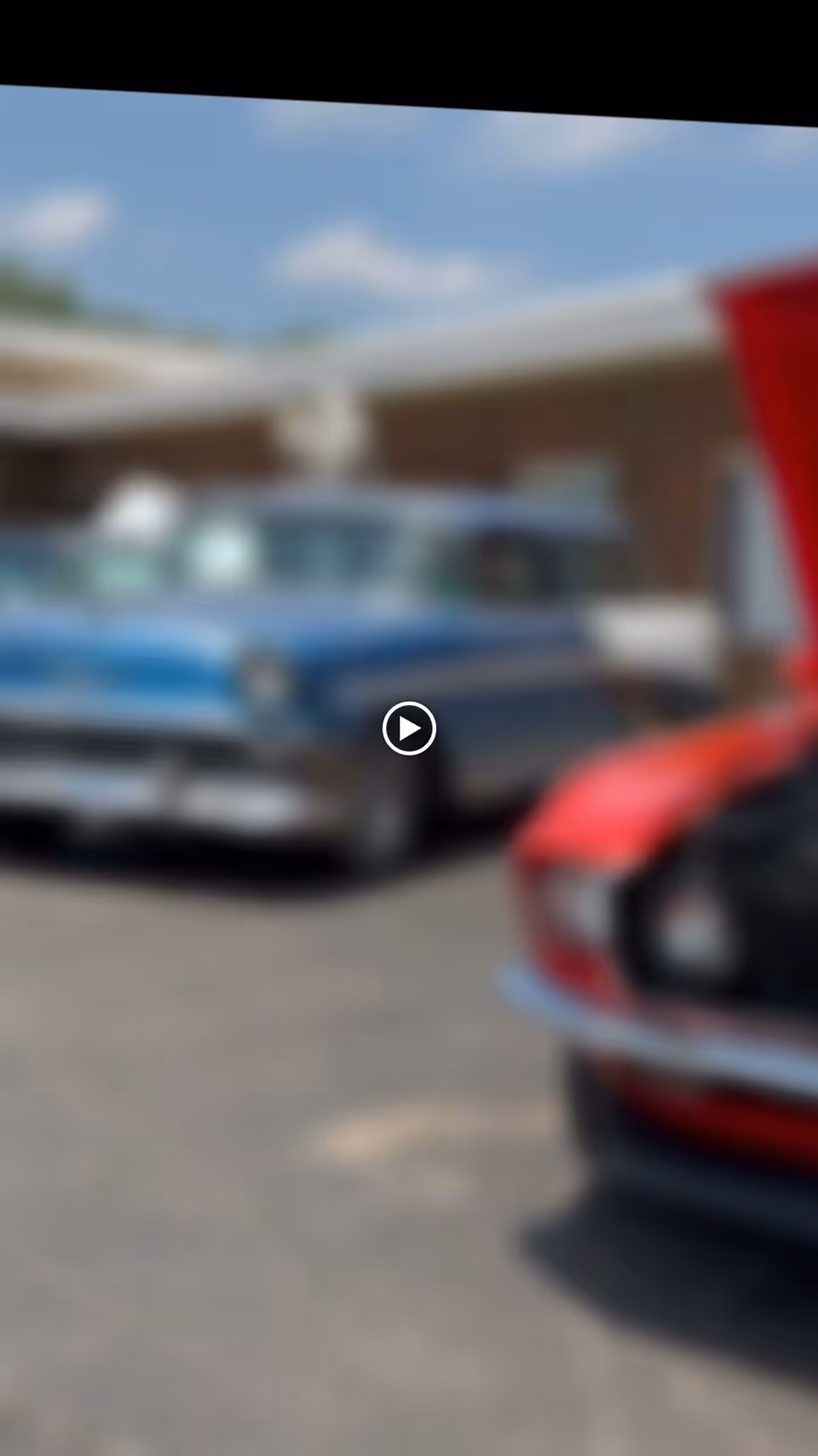 Blurred image showing two classic cars parked outside a building under a clear sky.