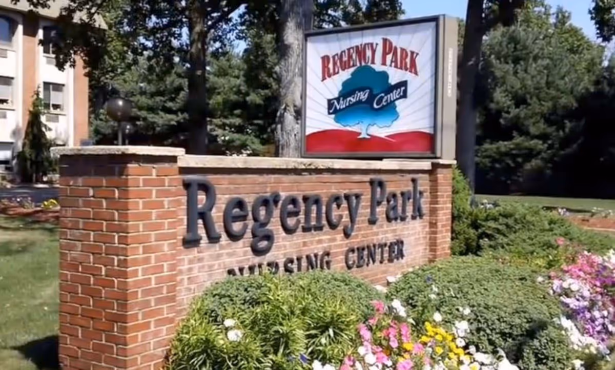 Outdoor view of a brick sign for Regency Park Nursing Center surrounded by green bushes and colorful flowers, with trees and part of a building visible in the background.