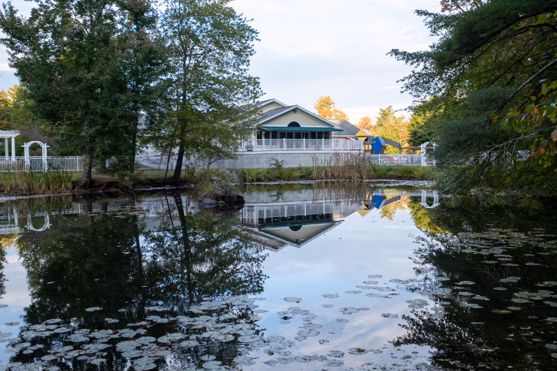 A serene pond with lily pads reflecting trees and a white building with a porch and a playground in the background, surrounded by greenery under a partly cloudy sky.