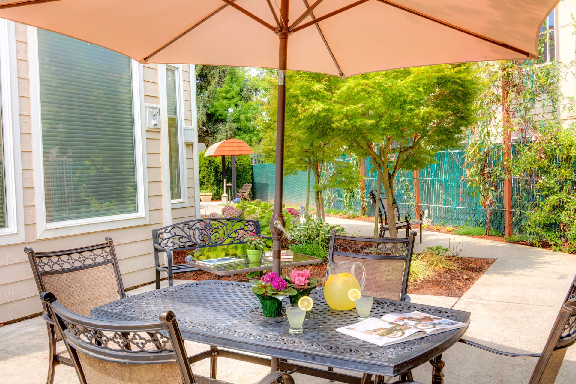 Outdoor patio with a table and chairs under a large umbrella, set with a pitcher of lemonade, glasses and flowers beside a landscaped walkway.
