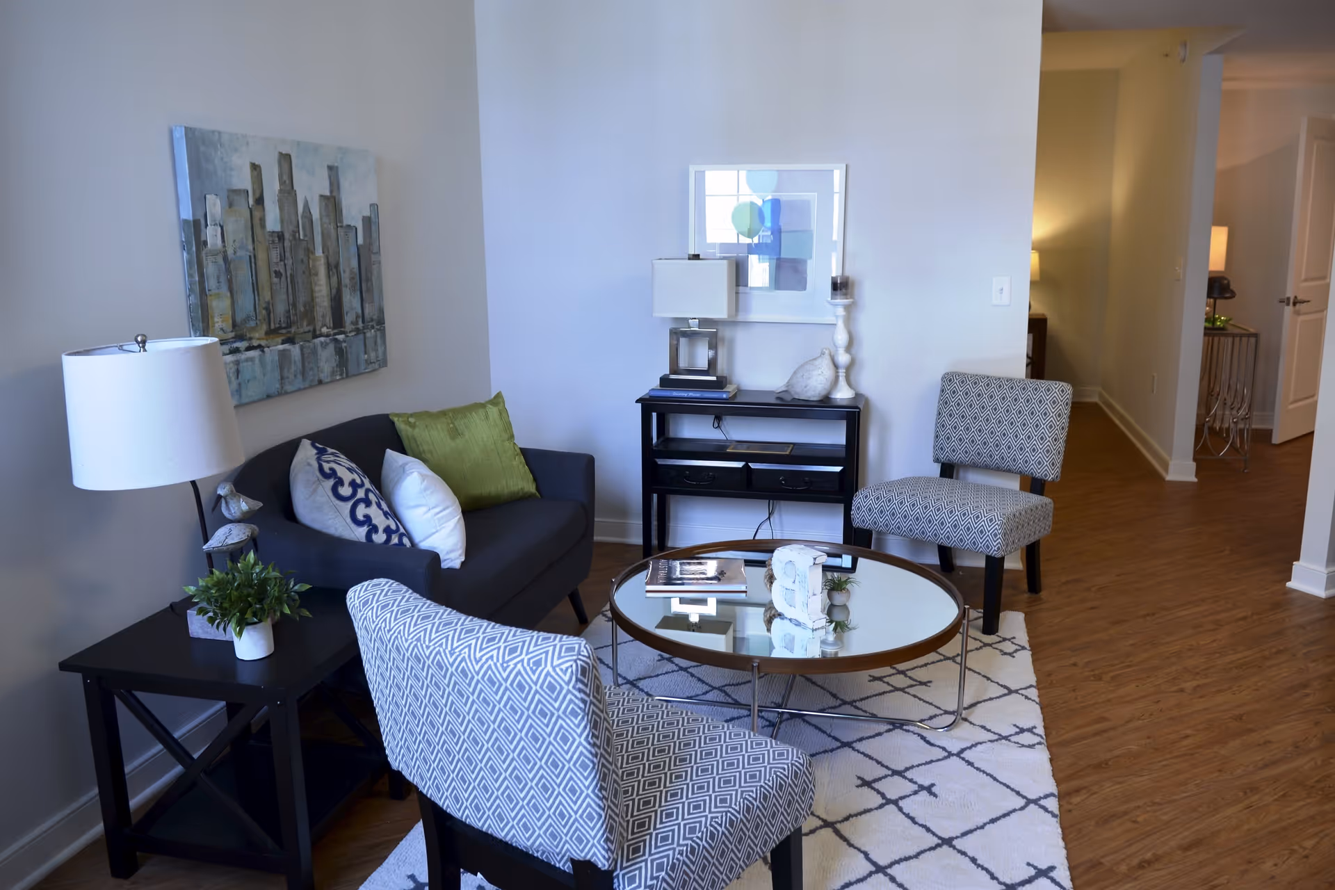 Bright furnished living room with a sofa, two patterned chairs, a round glass coffee table, side tables, lamps and wall art.