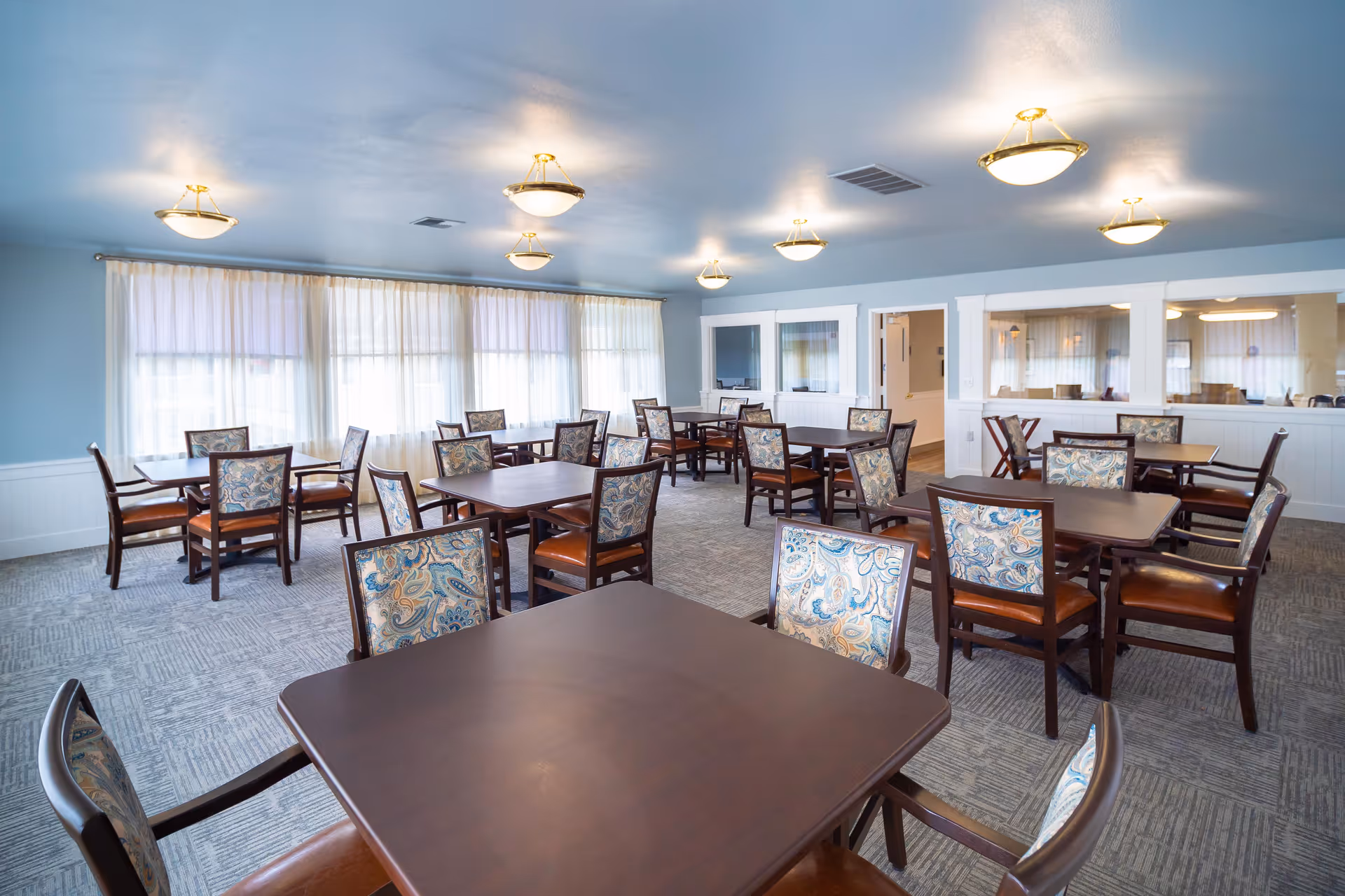 A bright dining room with multiple square wooden tables and chairs featuring patterned upholstery. The room has light blue walls, large windows with sheer white curtains, and ceiling lights with brass fixtures.