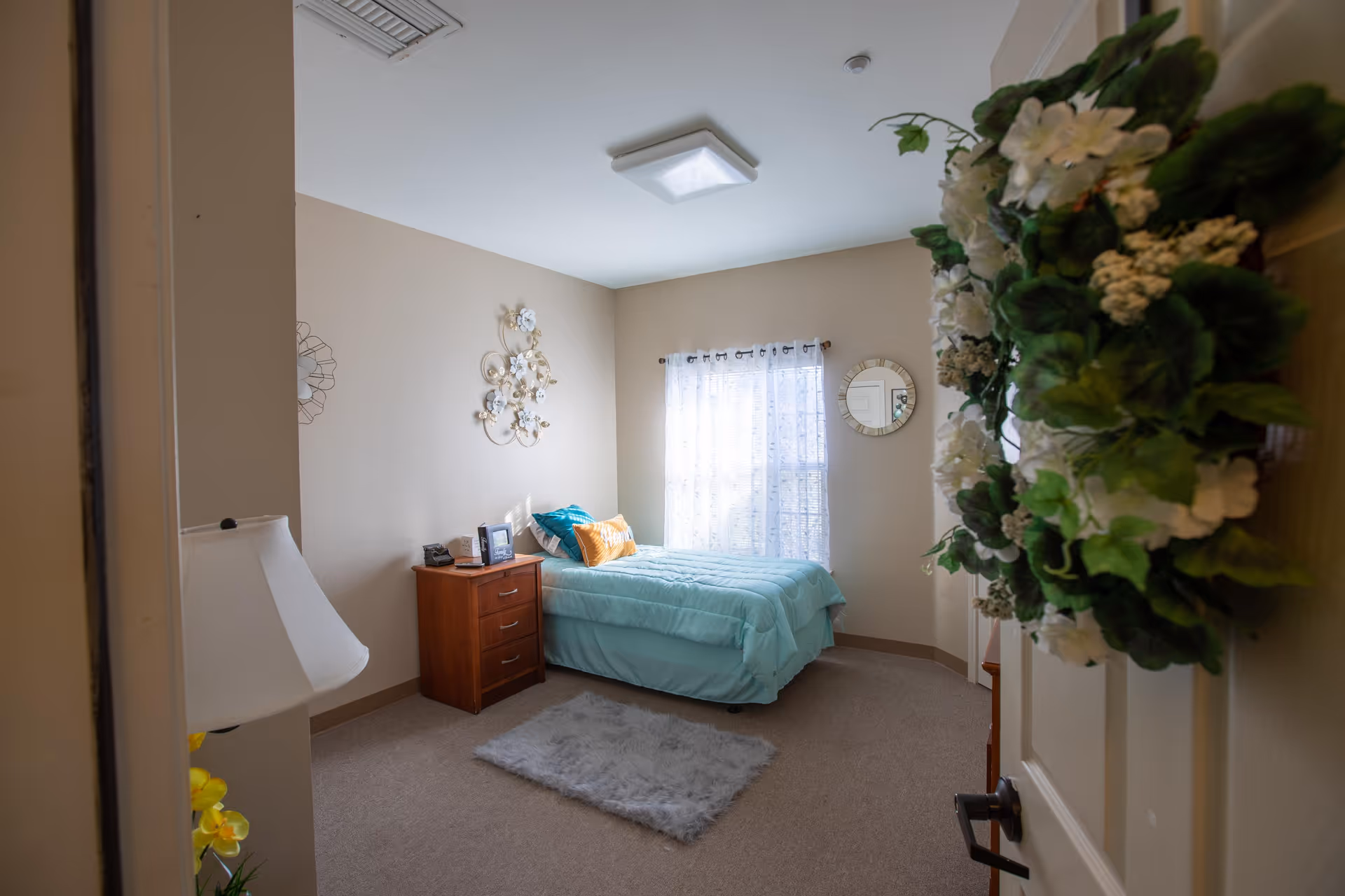 Small, well-lit bedroom with a twin bed in turquoise bedding, a wooden nightstand, wall decor, and a floral wreath on the door.