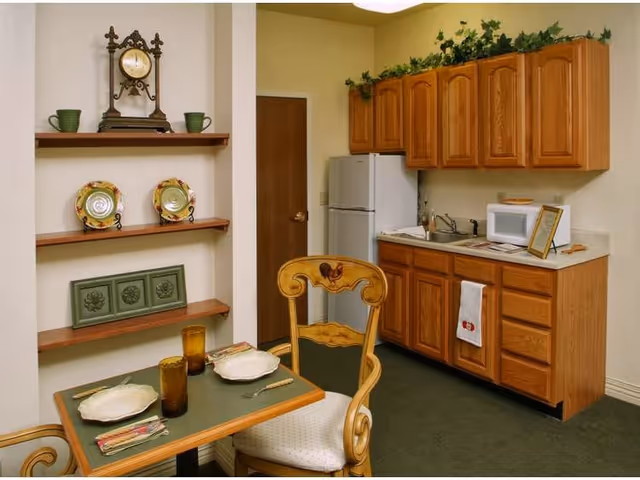 A small kitchen and dining area with wooden cabinets, a white refrigerator, microwave, and a sink. A table set for two with plates, utensils, and amber-colored glasses is in the foreground. Decorative plates and a clock are displayed on wall shelves.