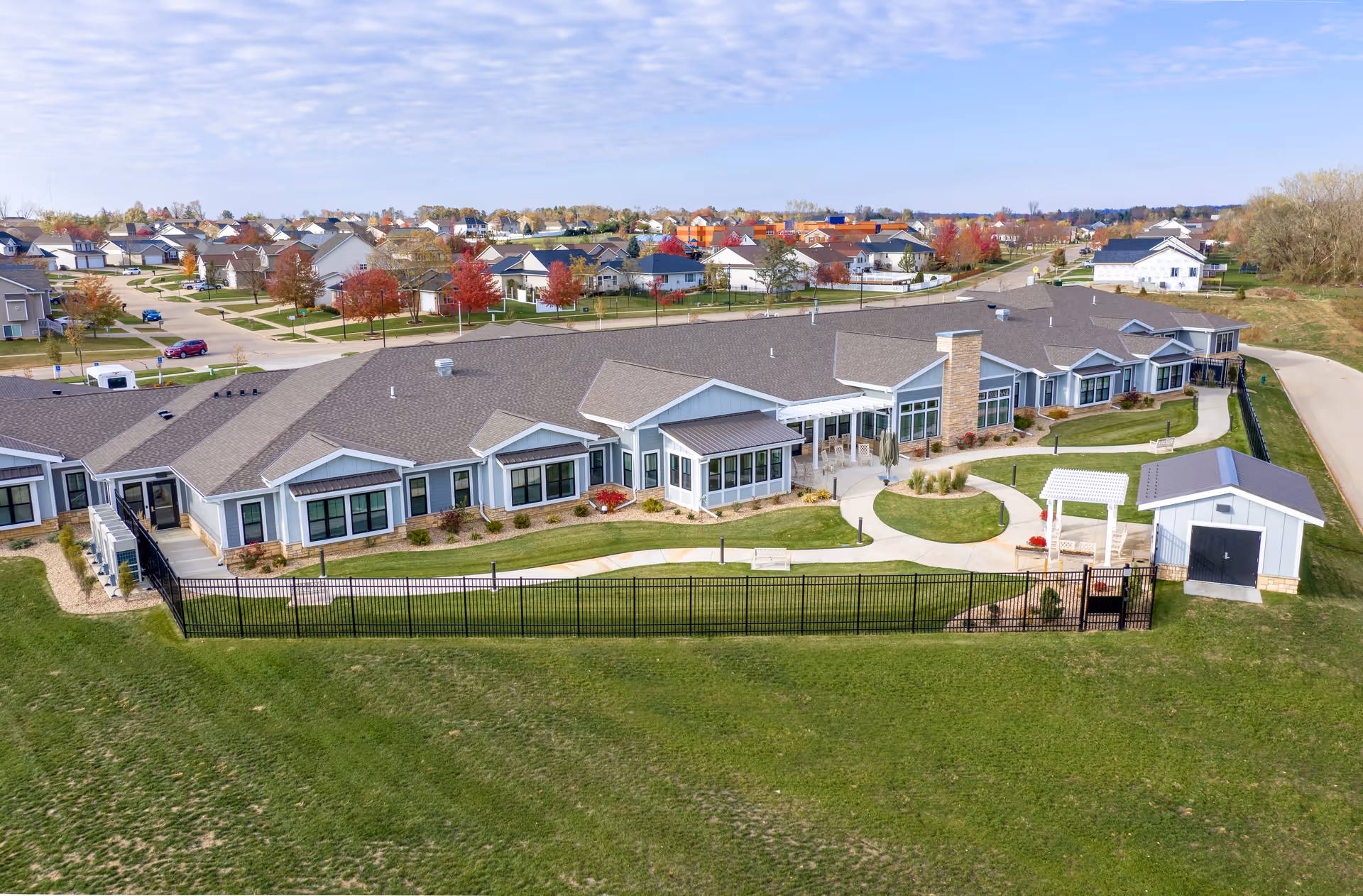 Aerial view of a single-story senior living facility named CountryHouse Residence, surrounded by a black fence with a small garden area and pathways. The building has a gray roof and light-colored exterior walls with large windows. The facility is located in a suburban neighborhood with houses and trees visible in the background under a partly cloudy sky.