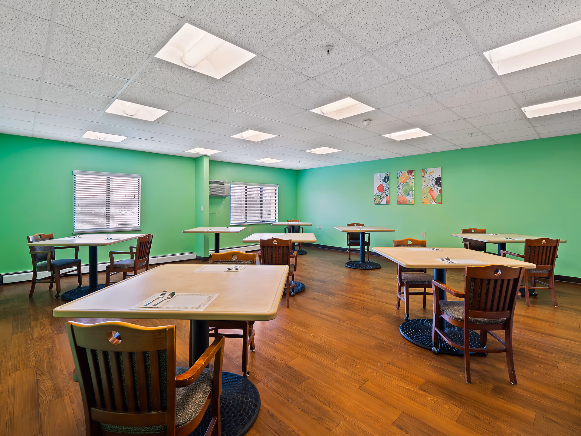 A dining room with green walls, wooden flooring, and several square tables each set with placemats, forks, and spoons. Wooden chairs with cushioned seats are arranged around the tables. Two windows with blinds and an air conditioning unit are visible on one wall, and three colorful fruit-themed paintings hang on another wall.