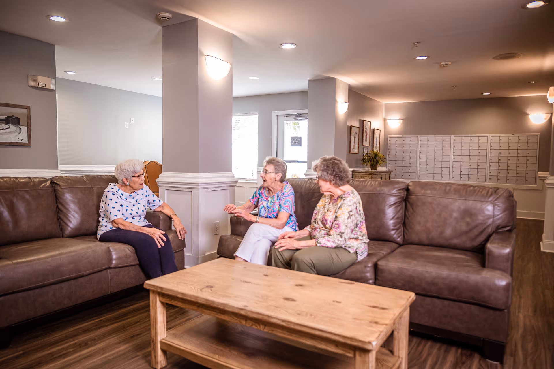 Three elderly women sitting on brown leather sofas in a well-lit common area with wooden flooring, a wooden coffee table, and a wall of mailboxes in the background.