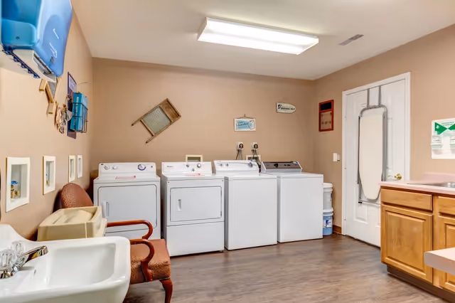 Interior laundry room with multiple washers and dryers, a sink, folding counter, chairs, and an ironing board on the door.
