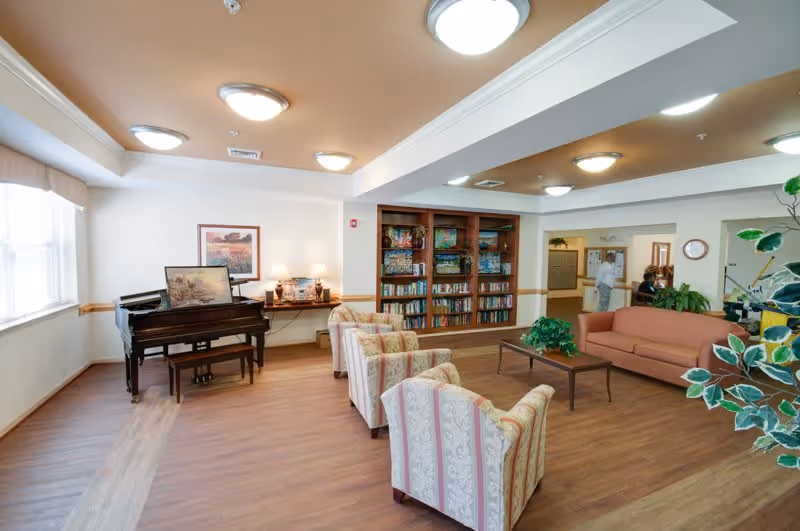 A bright and spacious living room area in a senior living facility featuring a grand piano, a wooden coffee table, a peach-colored sofa, and several patterned armchairs. There is a large bookshelf filled with books and games, a desk with lamps and framed pictures, and large windows letting in natural light. Two people are visible in the background near a hallway.