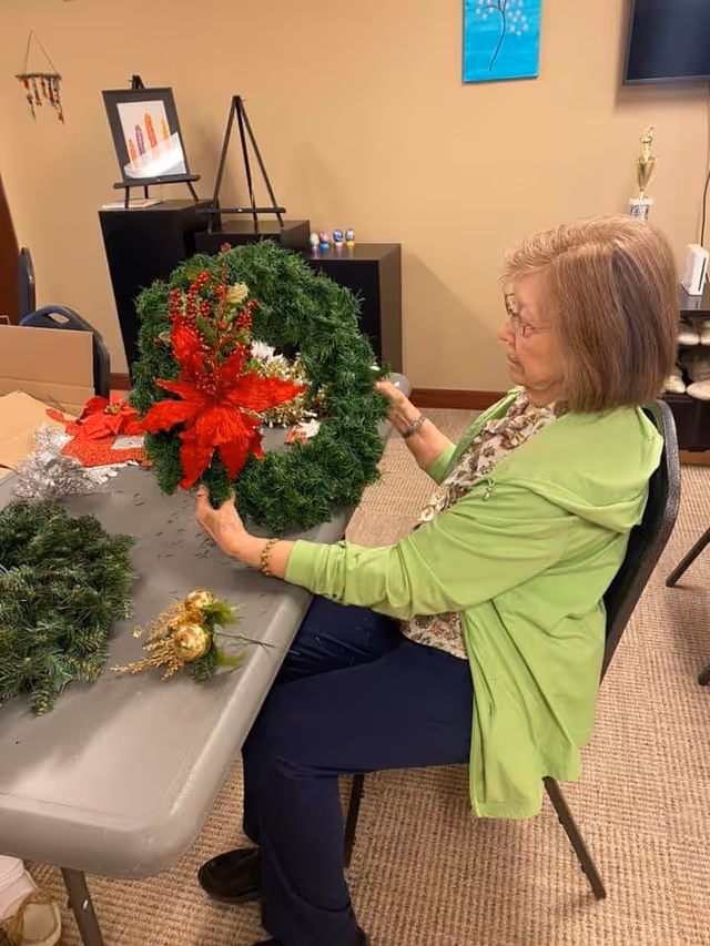 An elderly woman sitting at a table indoors, holding and examining a decorated Christmas wreath with red flowers and berries. The table has other wreath-making materials and decorations. The room has beige walls, a carpeted floor, and some furniture including a black shelving unit with framed pictures and small decorative items.