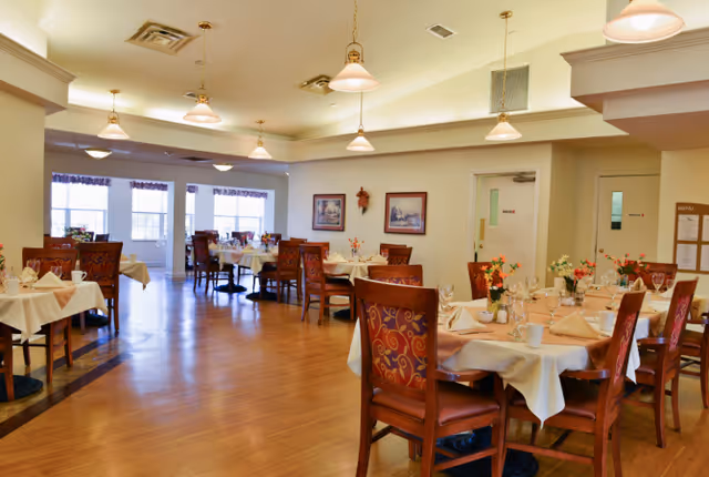 A spacious dining room with wooden floors and multiple tables covered with white tablecloths. Each table is set with napkins, glasses, and floral centerpieces. The room is well-lit with hanging pendant lights and has large windows with curtains allowing natural light to enter. There are framed pictures on the walls and a door in the background.