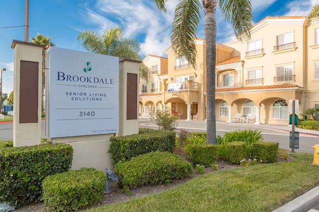 Exterior view of the Brookdale Carlsbad senior living facility with a large sign in the foreground displaying the facility name and address. The building is a multi-story structure with balconies, arches, and palm trees in the landscaped area around it under a blue sky with some clouds.