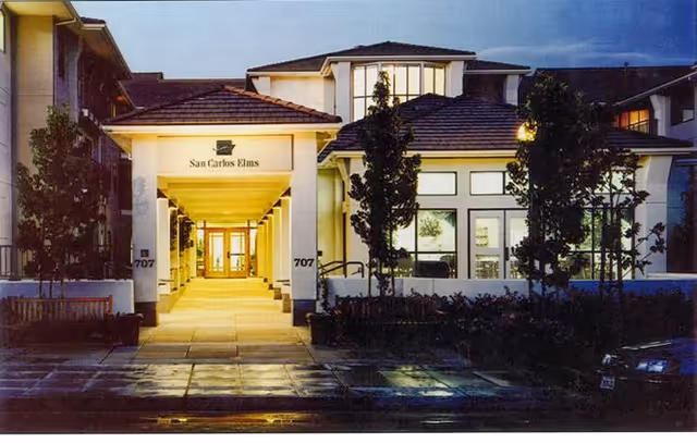Exterior front entrance of San Carlos Elms facility at dusk, featuring a well-lit covered walkway, trees, benches, and large windows.