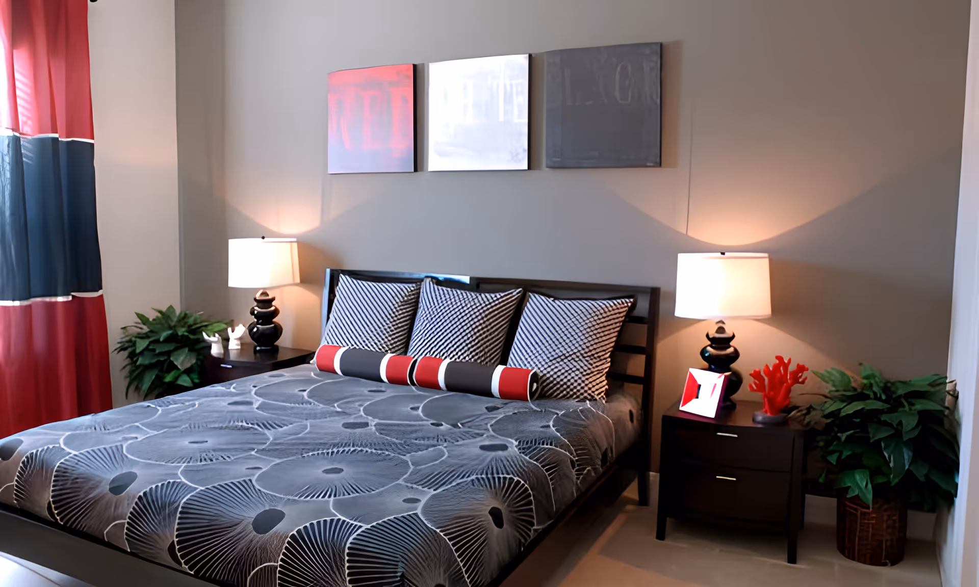 A neatly made bed with a black and gray floral patterned bedspread, three black and white striped pillows, and a cylindrical pillow with red, black, and white stripes. The bed is flanked by two dark wooden nightstands, each with a lamp and decorative items. There are two green potted plants on either side of the bed and three abstract art pieces hanging on the beige wall above the bed. Red and blue curtains cover the window on the left side of the room.