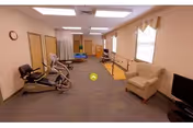 A rehabilitation room with exercise machines, parallel bars, chairs, and a TV under fluorescent lighting.