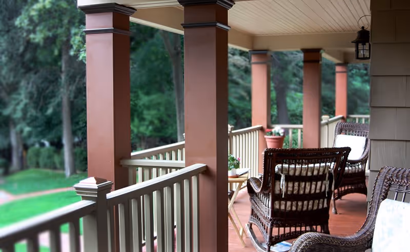 A covered outdoor porch area with brown wooden pillars and railing, featuring wicker rocking chairs with cushions and small potted plants on a wooden floor, overlooking a green garden with trees.