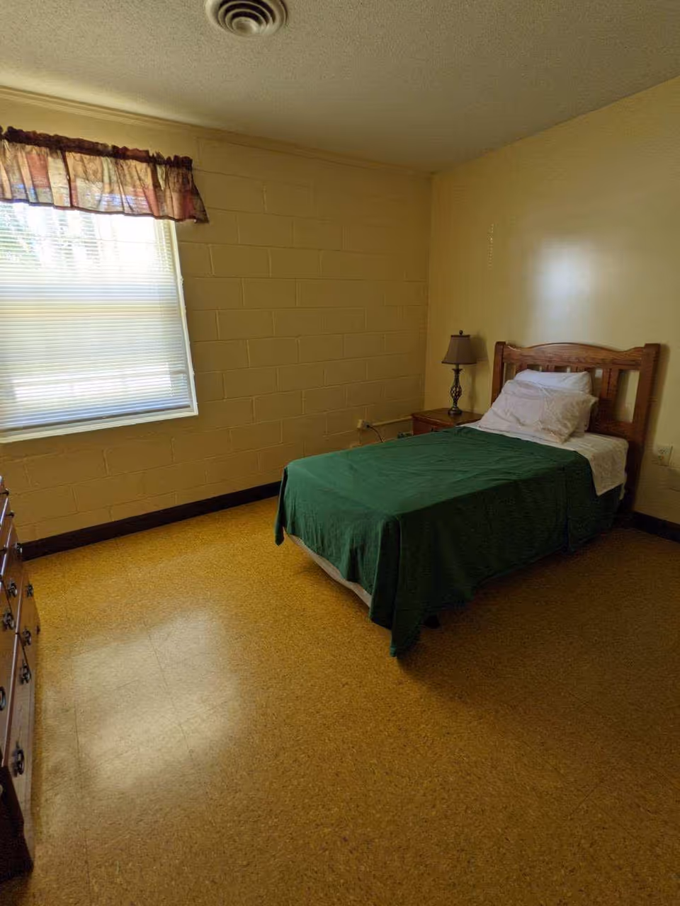A simple bedroom with a single wooden bed covered with a green blanket and white pillow. There is a wooden nightstand with a lamp next to the bed, a window with blinds and a valance curtain, and a wooden dresser on the left side. The walls are painted beige and the floor has a yellowish linoleum covering.