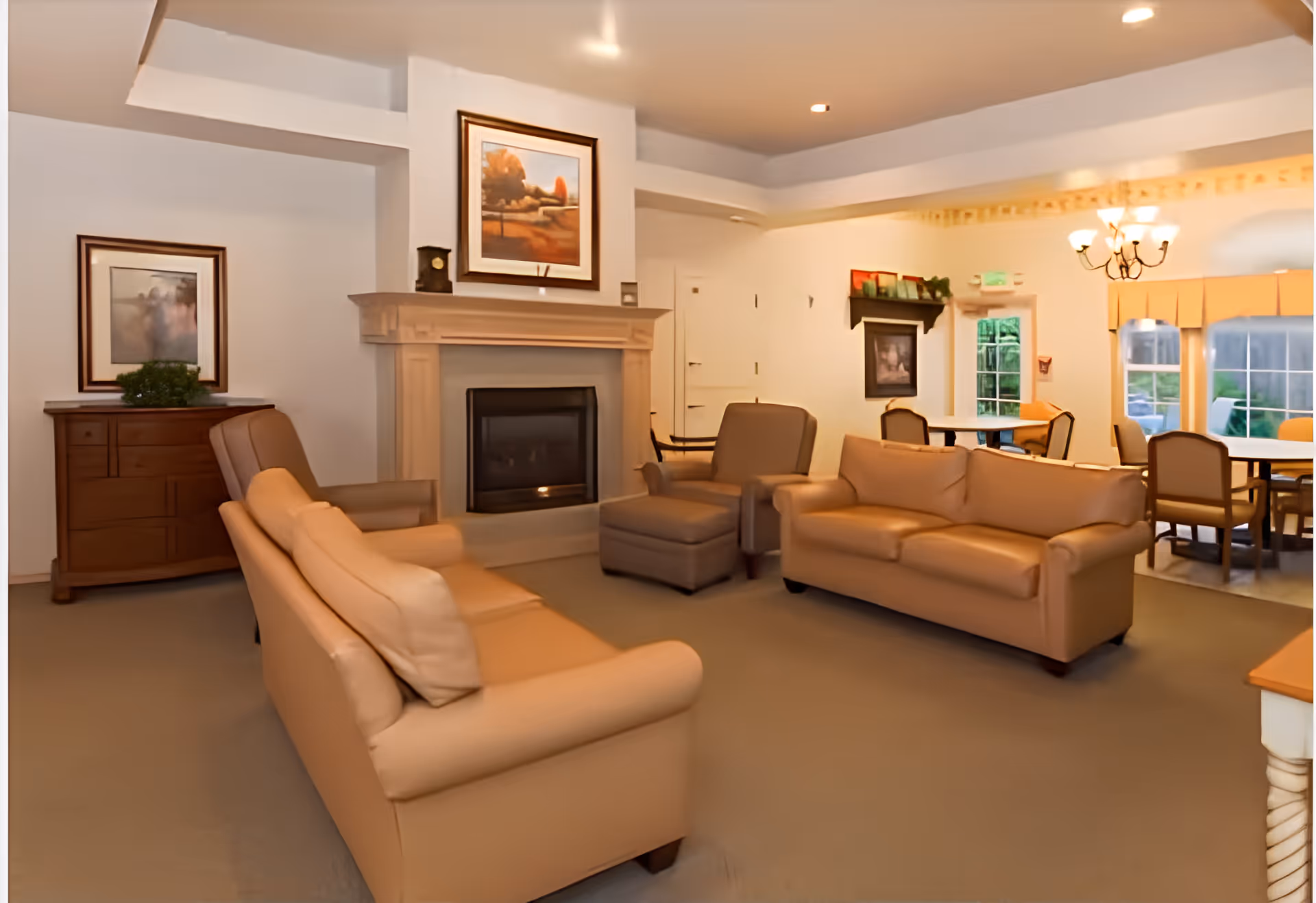 A cozy living room area with two beige leather sofas and a matching armchair arranged around a wooden fireplace. The room has light-colored walls adorned with framed artwork and a wooden chest of drawers with a plant on top. In the background, there is a dining area with tables and chairs near windows letting in natural light.