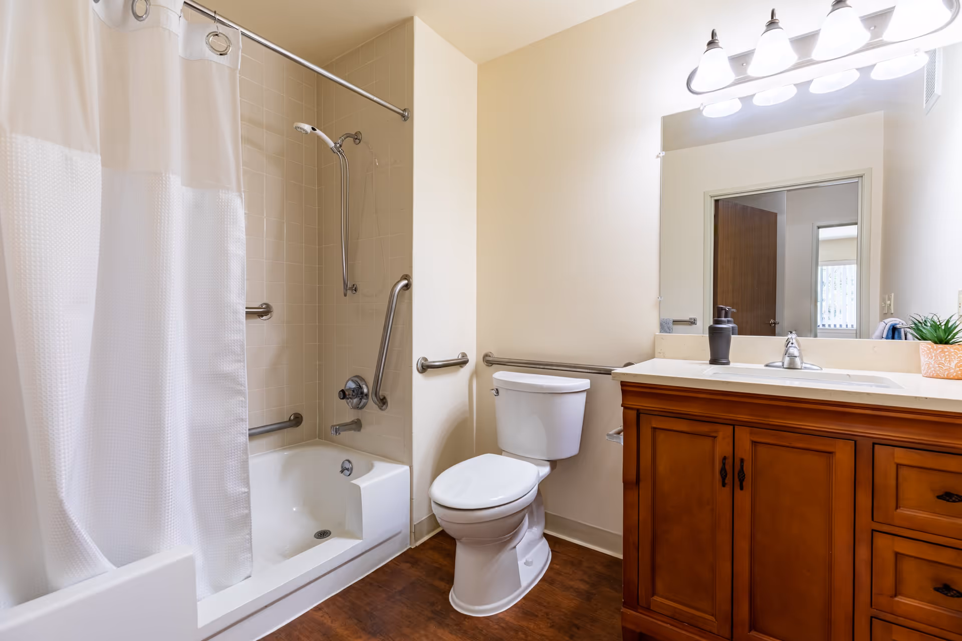 A clean and well-lit bathroom featuring a bathtub with a white shower curtain, a toilet with grab bars on the walls for accessibility, and a wooden vanity with a sink, soap dispenser, and a small potted plant. A large mirror and overhead lights are above the vanity.