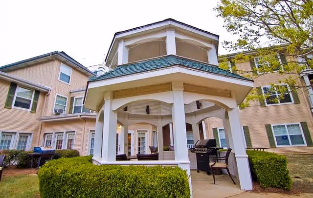 Outdoor covered gazebo area with seating and a grill, surrounded by trimmed bushes and part of a beige multi-story building with green shutters and windows in the background.