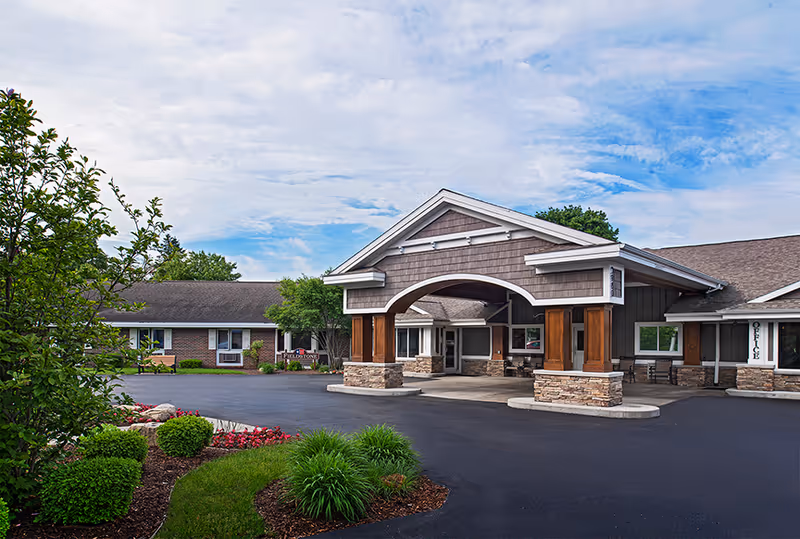 Exterior view of the American House Kentwood facility showing the main entrance with a covered driveway supported by stone and wooden pillars. The building has a combination of brick and siding with a well-maintained garden and shrubs in the foreground under a partly cloudy sky.