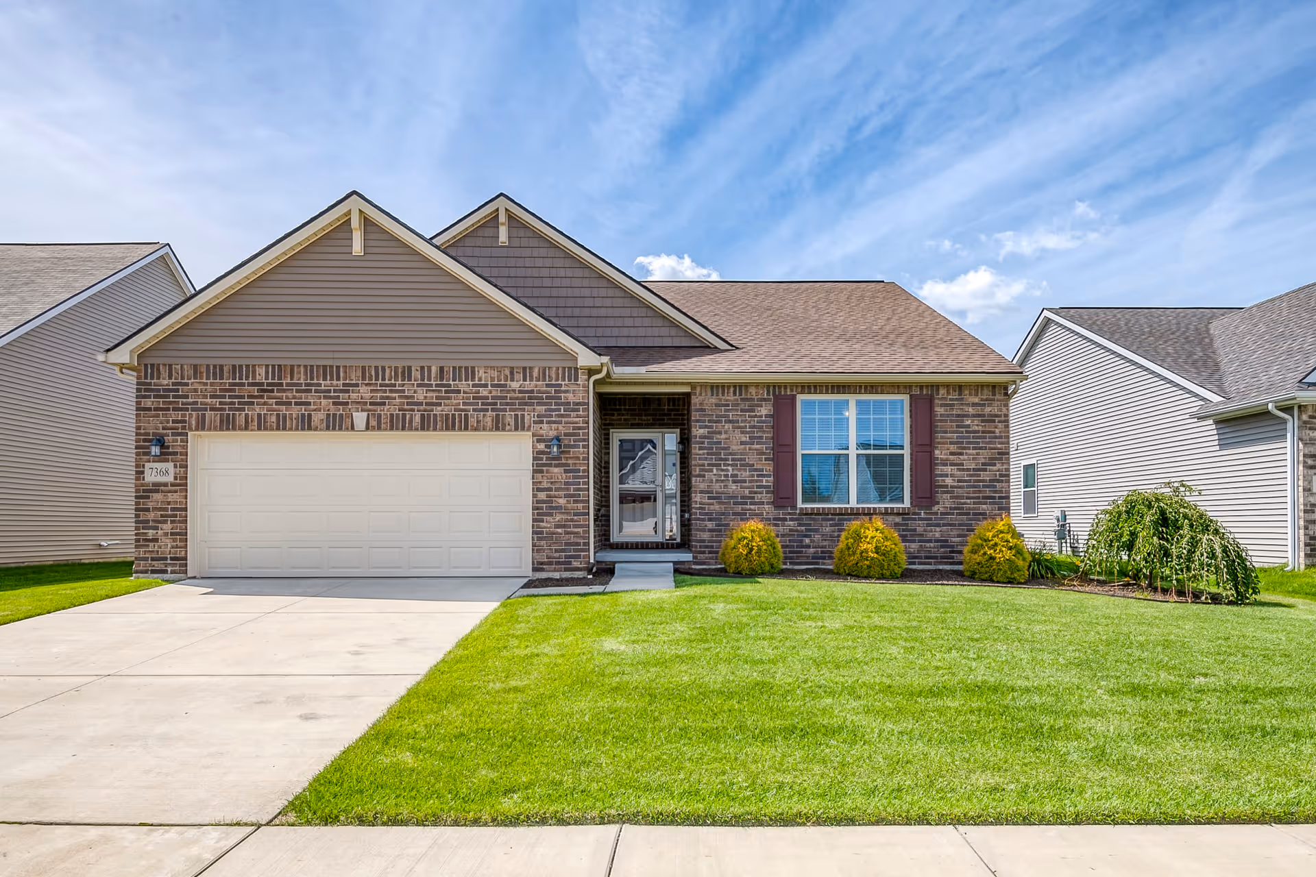 Front exterior view of a single-story brick house with a two-car garage, a concrete driveway, a green lawn, and a clear blue sky with some clouds.