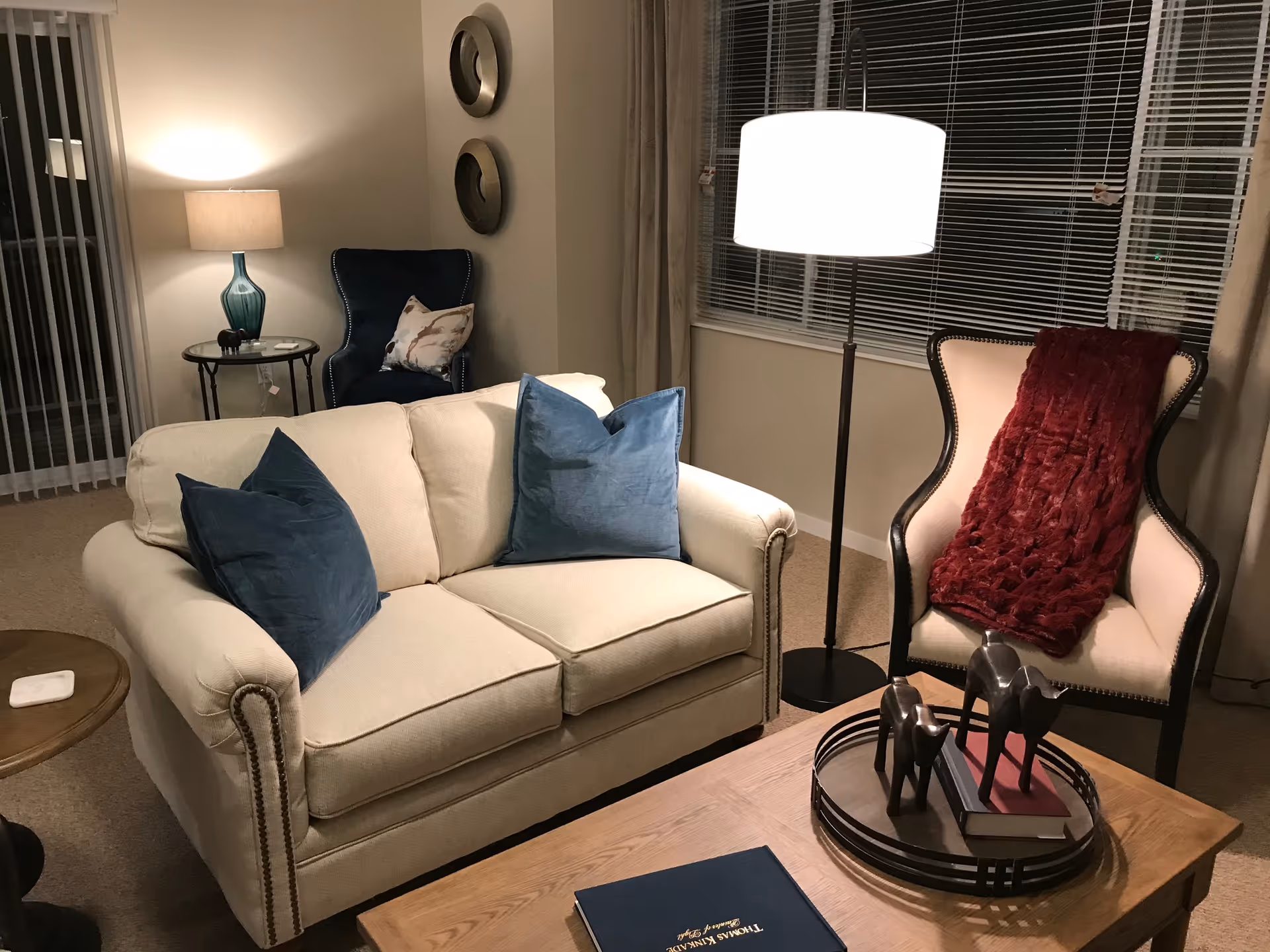 A cozy living room with a beige loveseat adorned with two blue throw pillows, a wooden coffee table with decorative items including a tray with two horse figurines and a book, a beige armchair with a red throw blanket, a standing lamp with a white shade, and a side table with a blue glass lamp and a small pillow on a dark blue chair in the corner.