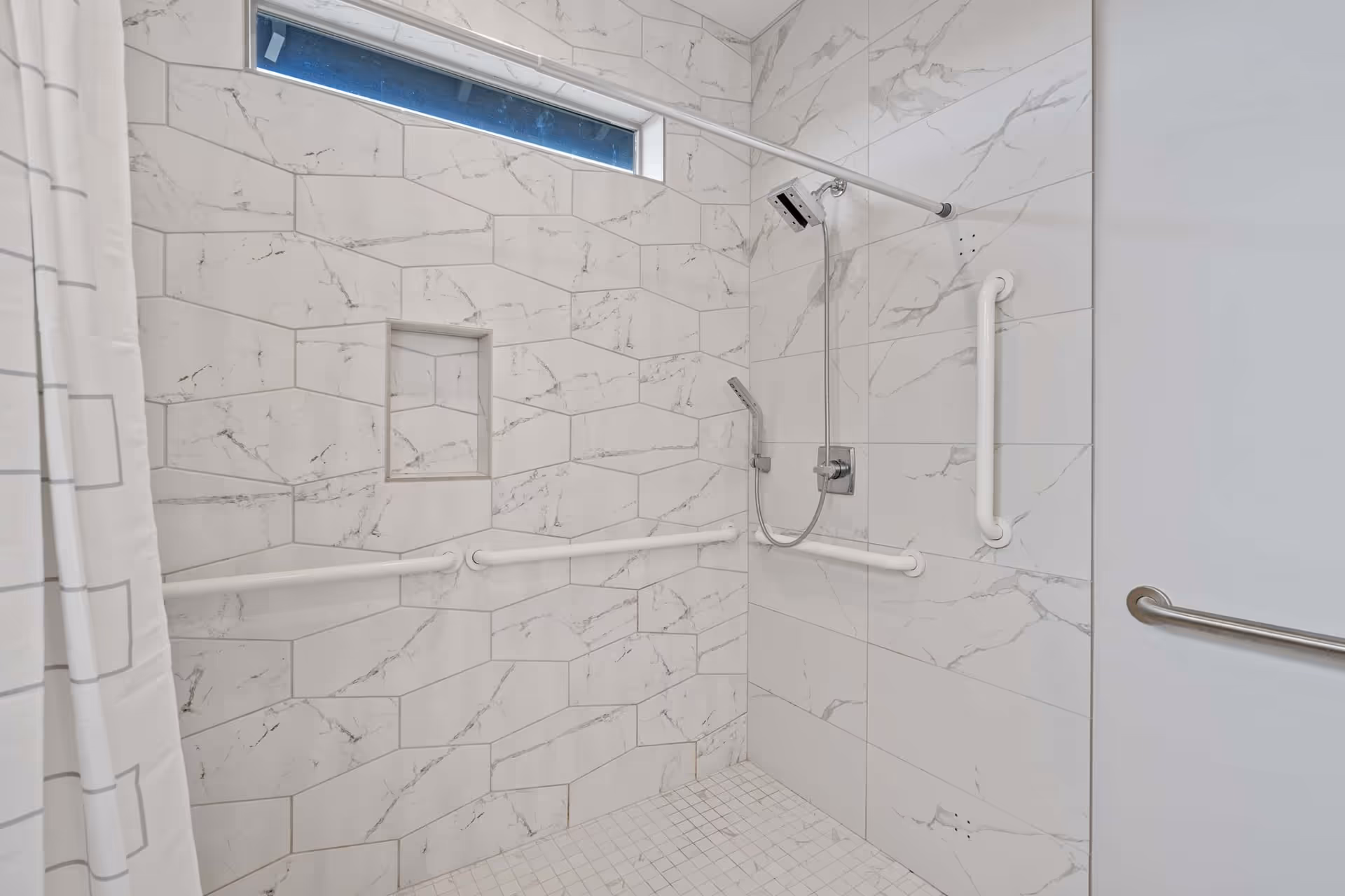 A modern accessible shower with white marble-patterned tiles on the walls and floor. The shower features multiple white grab bars, a handheld showerhead, a fixed square showerhead, a recessed shelf, and a white shower curtain with a geometric pattern. There is a narrow horizontal window near the ceiling.