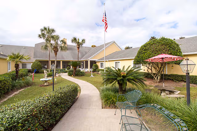 Outdoor courtyard area of a senior living facility with a curved concrete walkway, green bushes, palm trees, and a flagpole with an American flag. There are metal benches and a picnic table with a red and white striped umbrella. The building surrounding the courtyard is painted yellow with a gray roof.
