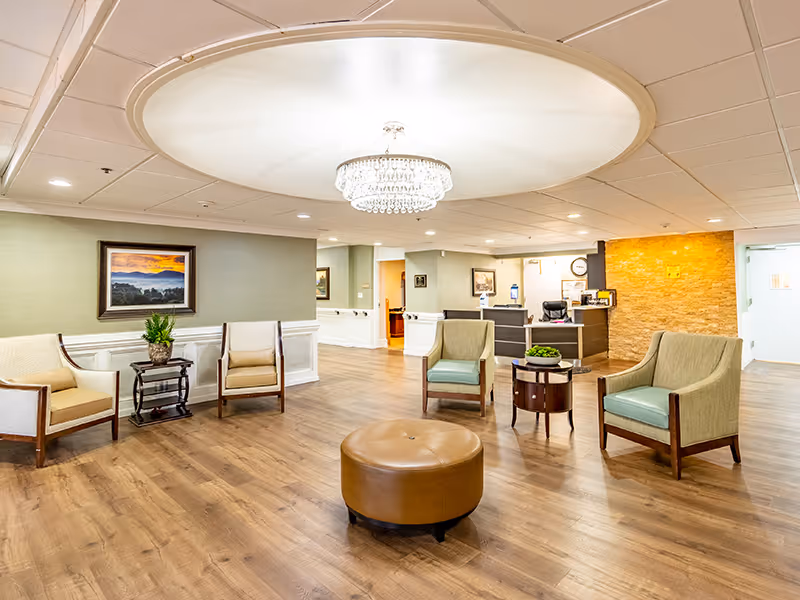 A spacious and well-lit senior living facility common area with wooden flooring, four cushioned armchairs arranged around a round leather ottoman, a small round table with a plant, a chandelier hanging from a recessed ceiling light, a reception desk in the background, and framed artwork on the walls.