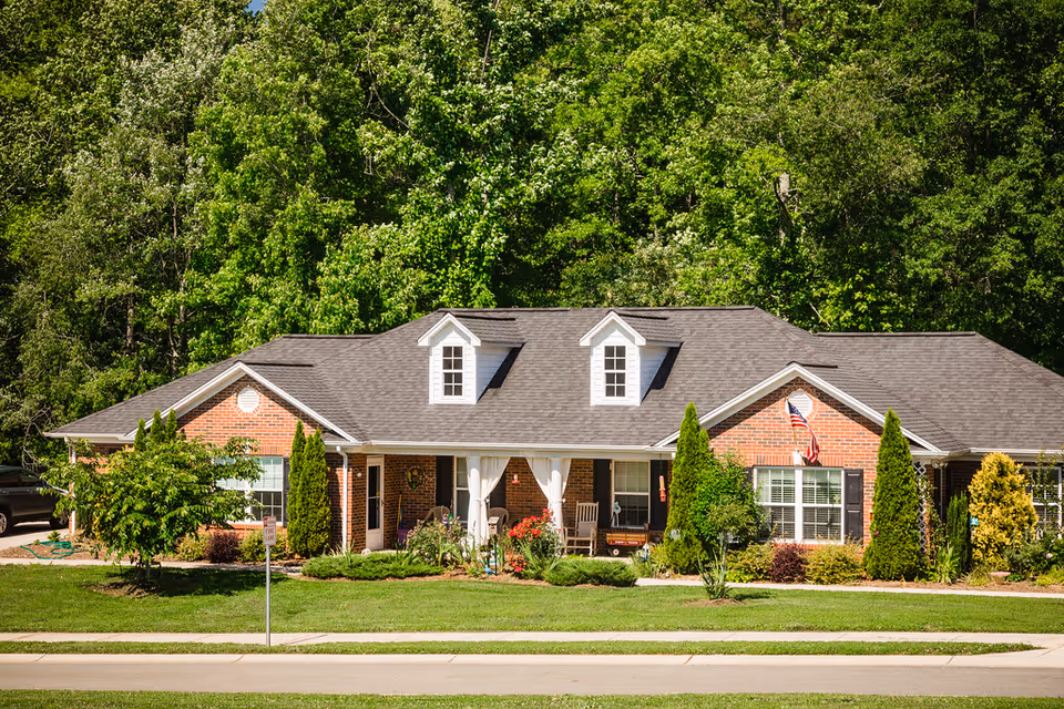 A single-story brick building with a gray shingled roof and two dormer windows. The building has a covered porch with white columns, rocking chairs, and various plants and shrubs in the front yard. There is an American flag mounted near one window, and tall green trees form a dense backdrop behind the building.