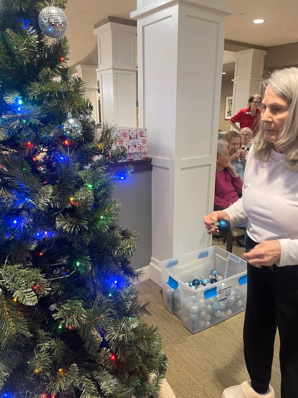 An elderly woman decorates a Christmas tree with blue ornaments in a well-lit indoor common area. In the background, several elderly people are seated and watching, with a staff member standing nearby. A large plastic bin filled with more blue and silver ornaments is on the floor next to the woman.