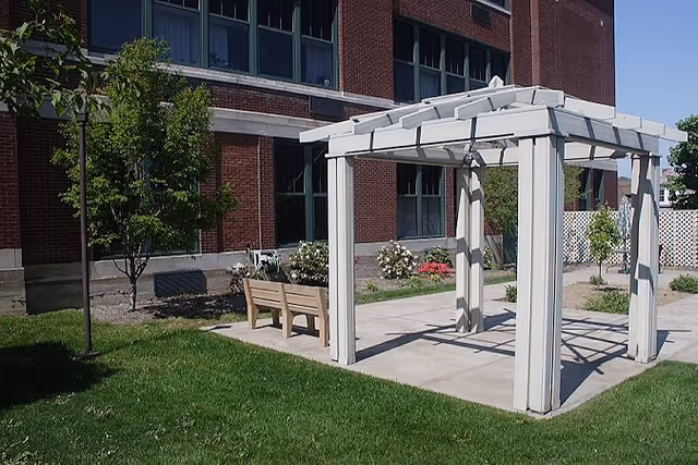 Outdoor area at Summit Retirement Residence featuring a white pergola on a concrete slab, a wooden bench, green grass, small trees, and a brick building with large windows in the background.