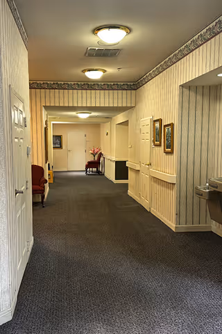 A carpeted hallway in a senior living facility with wallpapered walls featuring a floral border near the ceiling. The hallway is lit by ceiling lights and has framed pictures on the right wall. There are chairs placed along the walls and a water fountain on the right side near the foreground.