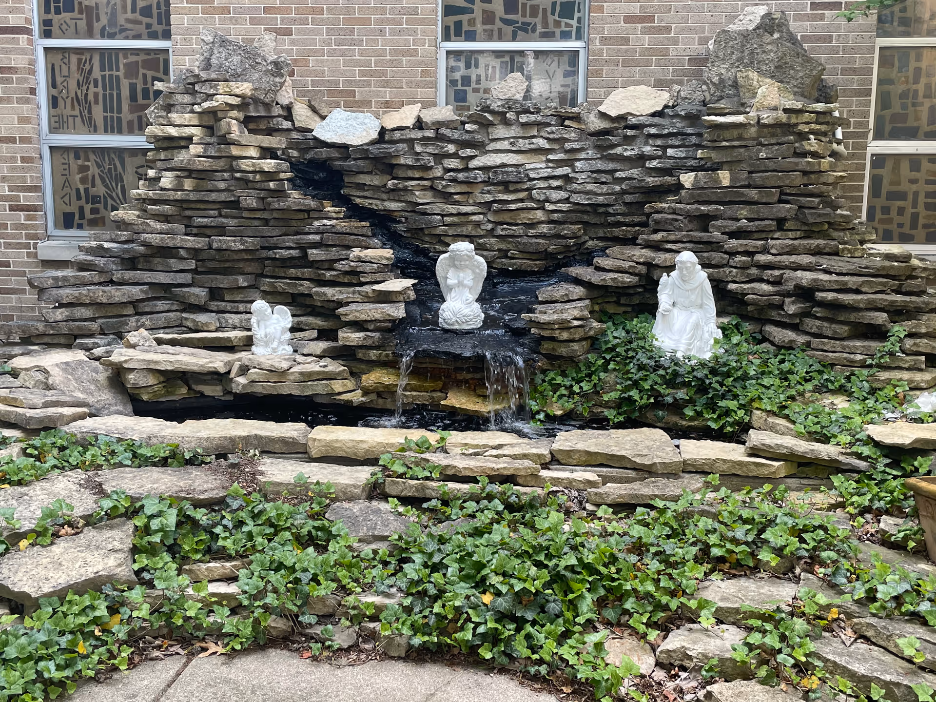 A stone water feature with a small waterfall surrounded by green ivy plants. Three white statues are placed on the rocks: one in the center, one on the left, and one on the right. The background shows a brick wall with windows featuring decorative glass.