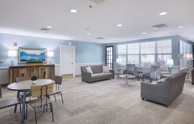 A spacious and well-lit living room area with light blue walls and beige carpet. The room features two gray sofas, two patterned armchairs, a round wooden table with four chairs, a wooden sideboard with a framed artwork above it, and several lamps providing soft lighting. Large windows with blinds allow natural light to fill the space.