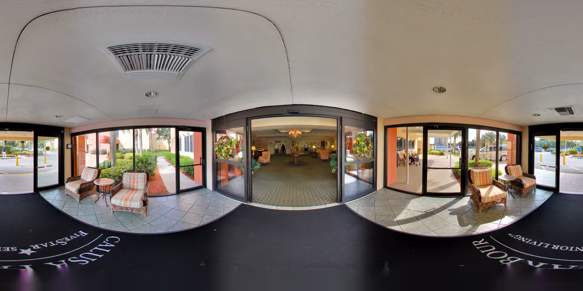 Panoramic view of the entrance lobby area of Calusa Harbour senior living facility, showing glass doors leading outside, two wicker chairs with plaid cushions and a small table on each side, and a glimpse into the interior common area with seating and plants.