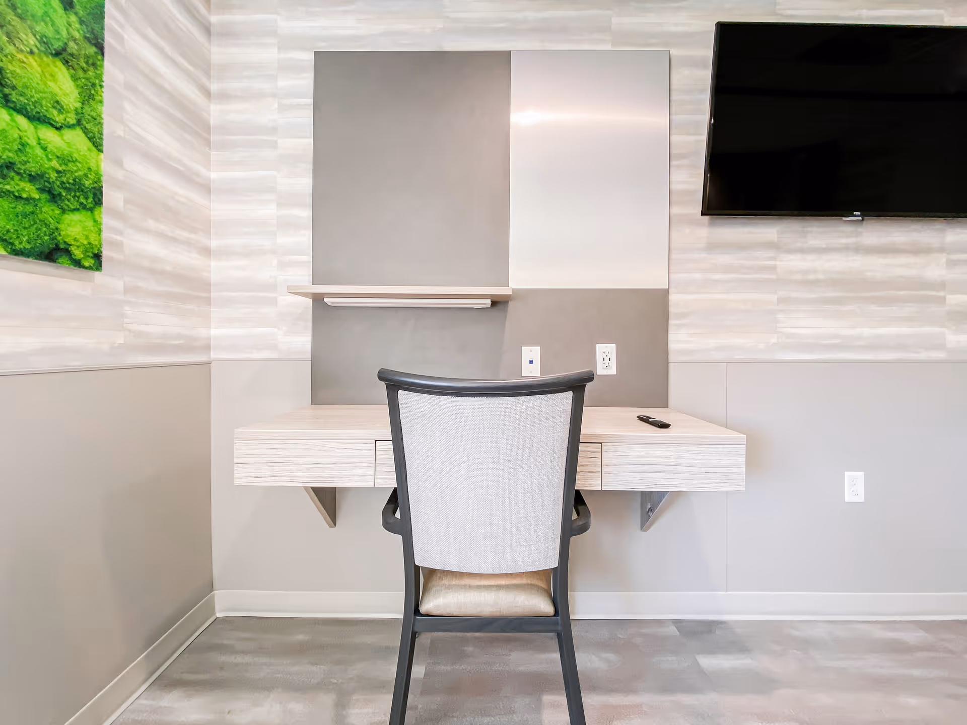A modern workspace with a light wood floating desk and a cushioned chair with a black frame. On the wall above the desk are two square panels, one gray and one white, and a small shelf. To the right, a flat-screen TV is mounted on the wall. The walls have a light gray and beige textured pattern, and a green moss-like wall art is partially visible on the left side.