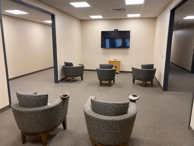 A small common area with six gray patterned armchairs arranged in two rows facing a wall-mounted flat screen TV. The room has beige walls, gray carpet, and fluorescent ceiling lights. There are two open doorways visible on the left and right sides of the room.