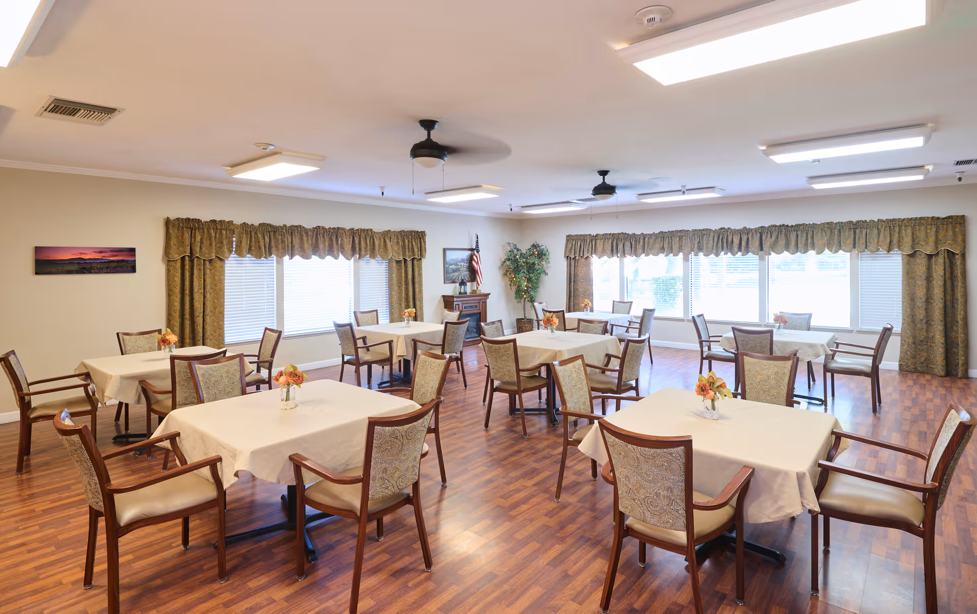 A bright and spacious dining room with several square tables covered with white tablecloths, each surrounded by four wooden chairs with cushioned seats and backs. The room has large windows with patterned curtains, wooden flooring, ceiling fans, and fluorescent lighting. Small flower arrangements are placed on each table, and there is an American flag and a potted plant in the corner.