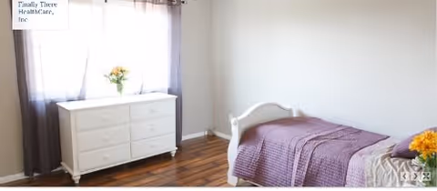 A simple bedroom with a single white bed covered with a purple quilt and a pillow. Next to the bed is a white dresser with six drawers, topped with a vase of yellow flowers. The room has wooden flooring and a window with sheer curtains allowing natural light to enter.