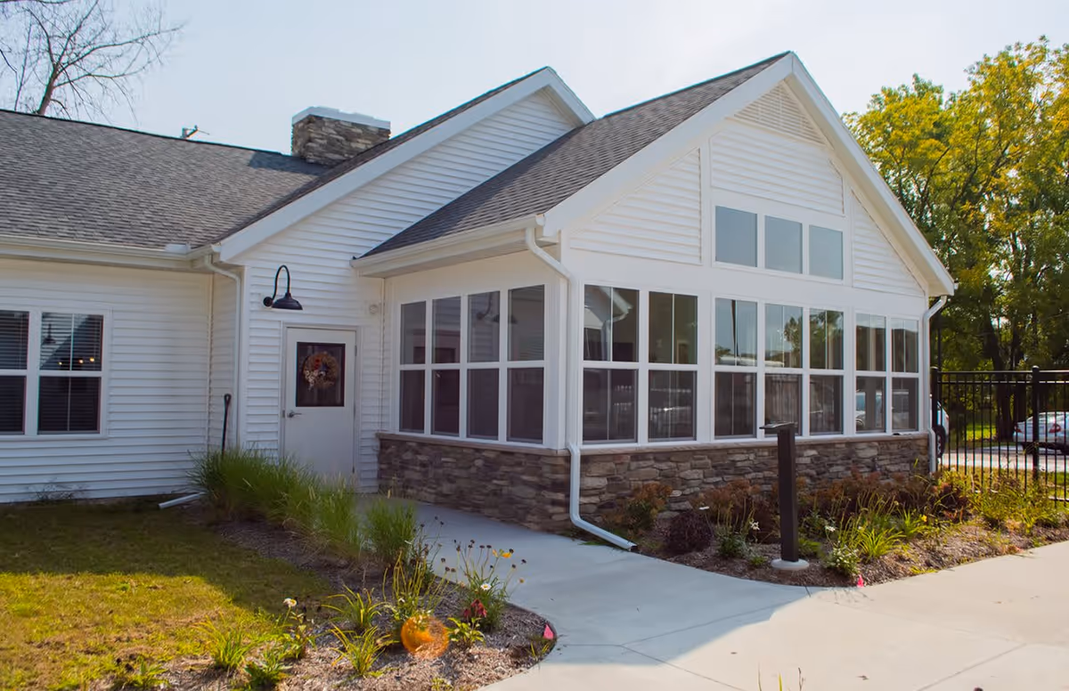 Exterior view of a white building with stone accents at the base, large windows, a door with a wreath, and a landscaped garden with grass and plants along a concrete walkway under a clear sky.